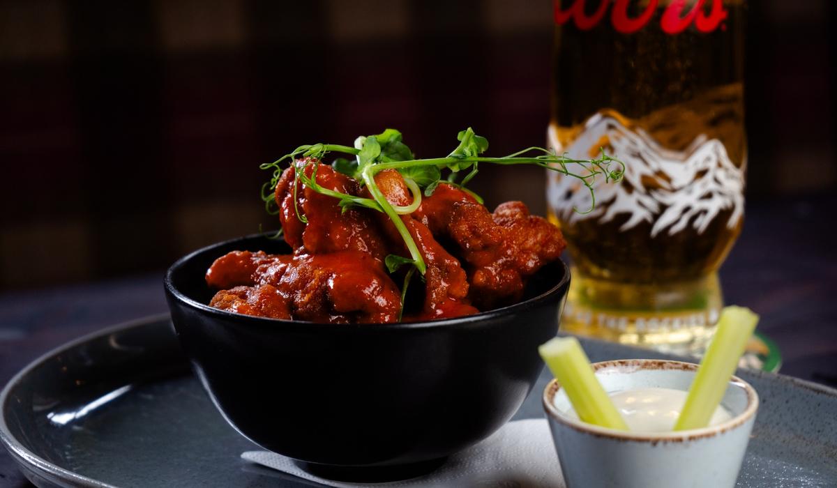 Crispy chicken wings coated in sauce served with celery and dipping sauce, with a pint of beer in the background. 