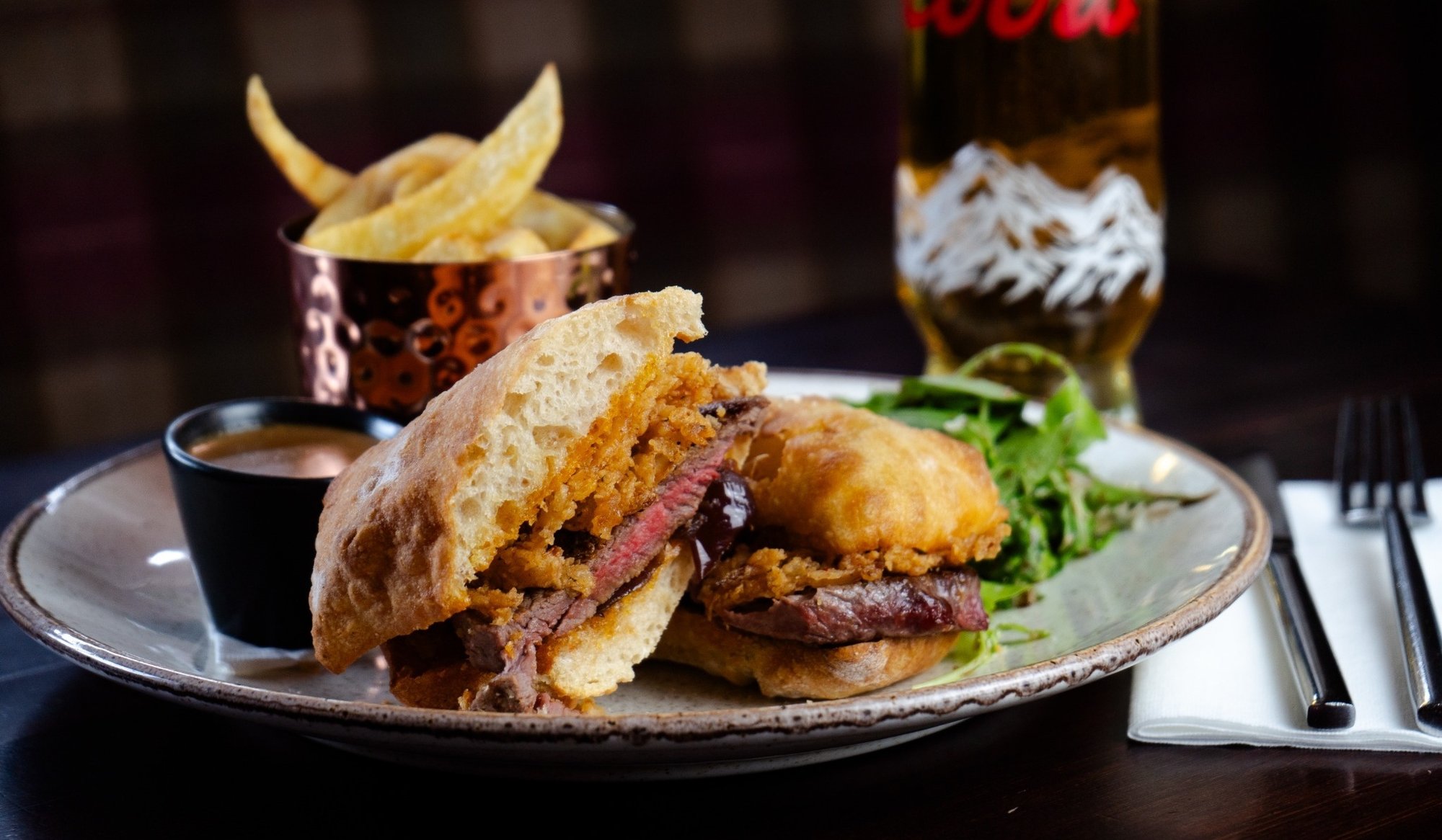 Steak sandwich with crispy onions served on a toasted bun, with chips, salad and dipping sauce, alongside a pint of beer.