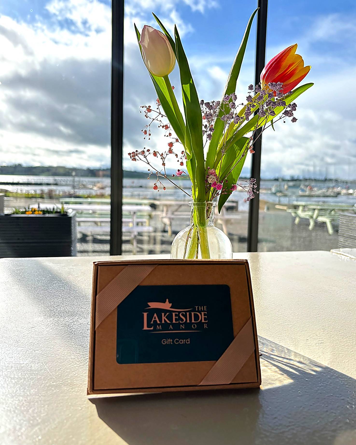 Lakeside Manor gift card displayed on a table beside a vase of tulips, with large windows overlooking the marina and lake in the background.